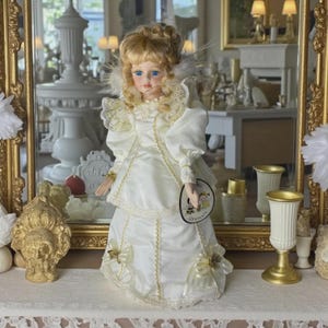 May include: A cream-coloured porcelain doll with blonde curly hair and blue eyes, wearing a dress with gold trim. The doll has a tag attached. It is displayed in front of a mirror.