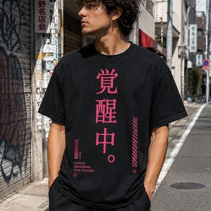 以下が含まれることがあります： ピンク色の日本語の文字と「SYSTEM AWAKENING. STAY FOCUSED」の文字が入った黒いTシャツ。Tシャツはクルーネックで半袖です。人物は街の通りに立っています。