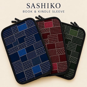Könnte beinhalten: Drei rechteckige Reißverschlusshüllen für Bücher und Kindles mit der Aufschrift SASHIKO BOOK & KINDLE SLEEVE. Jede Hülle zeigt ein Patchwork-Design mit weißen geometrischen Nähten in Blau, Dunkelrot und Dunkelgrün mit schwarzem Rand.