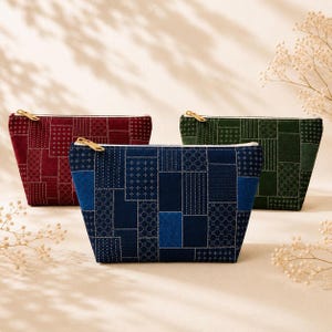 May include: Three zippered pouches in deep red, navy blue, and forest green. Each pouch features a patchwork design with various geometric patterns in white stitching. The pouches have gold-toned metal zippers along the top edge.