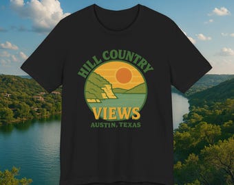 May include: Black t-shirt featuring a circular graphic with the text "HILL COUNTRY VIEWS" and "AUSTIN, TEXAS". The graphic depicts a landscape with a sunset, water, and cliffs. The shirt is made of a soft material.