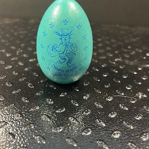 Puede incluir: Un huevo de Pascua de color turquesa con un diseño de conejo de dibujos animados y el texto "WHITE HOUSE EASTER EGG ROLL 2005". El huevo está decorado con estrellas y un conejo con una cesta.