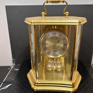 May include: A gold-toned mantel clock with a glass enclosure featuring etched rose designs on the side panels. The clock has a circular face with Roman numerals and a pendulum with four gold spheres. A metal ruler shows a width of 11 inches.