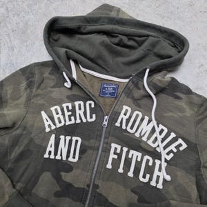 May include: A green camouflage zip-up hoodie with the text "Abercrombie and Fitch" in white block letters. The hoodie has a drawstring hood and a full-length zipper. The fabric appears to be a soft, worn material.