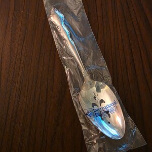 May include: A silver-toned serving spoon with an ornate handle design, encased in a clear plastic sleeve. The bowl of the spoon features a decorative cutout pattern and the printed text International DeepSilver in blue ink on the plastic surface.