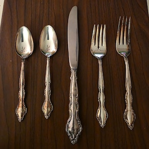 May include: A set of five pieces of silver-toned flatware arranged vertically, consisting of two spoons, one dinner knife, and two forks. Each piece features an ornate, embossed handle design with a polished finish and visible signs of tarnish.