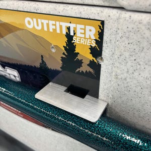 May include: A close-up of a product section with the text "OUTFTTER SERIES" on a yellow and blue background. The product has a teal and black speckled finish and a silver metal bracket.