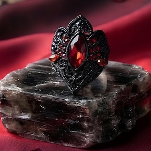 May include: A black metal ring featuring an ornate, filigree-style setting with a central marquise-cut blood red gemstone. The ring is displayed on a rectangular, translucent gray stone block. Text reads: Master Reiki Infused Blood Red Gemstone Amulet.