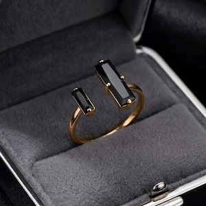May include: An open-style gold ring featuring two black rectangular baguette-cut stones. The larger stone is positioned at the top, while the smaller stone sits at the end of the open band. The ring has a polished gold finish and a sleek design.