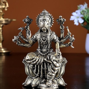 Può includere: Una statua color bronzo metallizzato della divinità Varaha, raffigurata con testa di cinghiale e braccia multiple. La figura è seduta a gambe incrociate su una base di loto stilizzata, adornata con gioielli complessi, collane e abiti dettagliati.