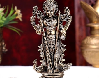 May include: A bronze-toned metal statue of a deity standing on a square base. The figure has four arms, holding a spear and a flag, and is adorned with intricate jewelry and a crown. A peacock is positioned at the feet of the standing figure.