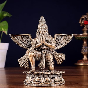 May include: A metallic bronze-toned statue of a winged deity kneeling on a lotus-shaped base. The figure has a pointed crown, detailed facial features, and hands pressed together in a prayer position, with large feathered wings extended outwards.