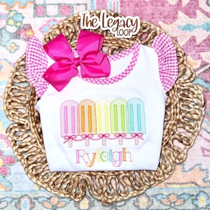 May include: White baby top with a colourful popsicle design and the name "Ryleigh" embroidered in rainbow colours. The top features pink gingham trim and a large pink bow. The Legacy Loop logo is visible.