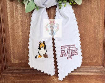 May include: A white fabric door hanger with scalloped edges featuring an embroidered tri-color collie dog with a football on the left tail and a maroon Texas A&M logo on the right tail, suspended from a wreath of white flowers and green foliage.