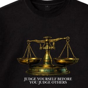 Könnte beinhalten: Schwarzes T-Shirt mit einer detaillierten Illustration einer goldenen Waage. Die Waage hat zwei Schalen, die an Ketten hängen. Darunter steht der Text "JUDGE YOURSELF BEFORE YOU JUDGE OTHERS."