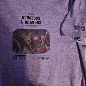 May include: Purple t-shirt featuring the text "TSR DUNGEONS & DRAGONS" above a fantasy illustration of a dragon and a figure in a dungeon. The sleeve has a small "D&D" logo. The text "FANTASY ADVENTURE GAME BASIC RULEBOOK" is also present.