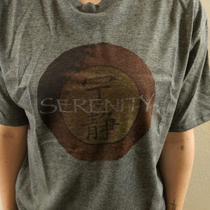 May include: A gray t-shirt featuring a circular design with the word "SERENITY" and Japanese characters. The design is in shades of brown and gold. The shirt has a crew neck and short sleeves.