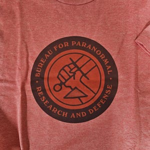 May include: Red t-shirt with a circular graphic. The graphic features the text "BUREAU FOR PARANORMAL RESEARCH AND DEFENSE" around a central design of a fist holding a weapon. The shirt has a crew neck and short sleeves.