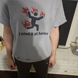 May include: Gray t-shirt with a graphic of a stick figure running with flames and the text "I tried it at home". The shirt is made of a soft material and has short sleeves. The person is wearing brown sweatpants.