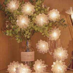 Pode incluir: Uma sequência de luzes decorativas com flores brancas e brilhantes. As luzes estão dispostas em torno de um vaso com vegetação e um copo de vinho, criando um ambiente suave e acolhedor. As flores têm uma aparência brilhante e texturizada.