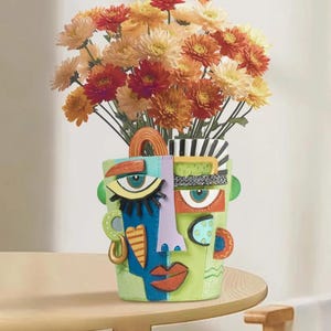 Könnte beinhalten: Eine dekorative, abstrakte Gesichtsvase in Grün, Blau und Orange, gefüllt mit orangefarbenen und gelben Blumen. Die Vase hat ein einzigartiges Design mit Gesichtsmerkmalen und geometrischen Formen, die auf einem Holztisch steht.