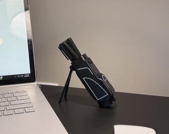 May include: A black pen holder with white accent lines, holding several black pens. The pen holder is on a stand, and is on a black desk next to a laptop. The laptop screen displays a spreadsheet.