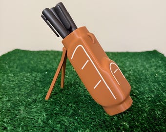 May include: A brown golf bag-shaped pen holder with white accent lines, holding several black pens. The holder is propped up on a stand, set against a backdrop of green turf.