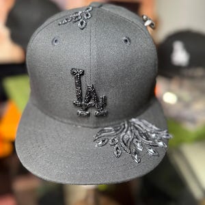 May include: A gray baseball cap with the letters "LA" embellished with black rhinestones. The cap features decorative rhinestone accents on the crown and brim, adding a touch of sparkle to the design.