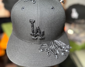 May include: A gray baseball cap with the letters "LA" embellished with black rhinestones. The cap features decorative rhinestone accents on the crown and brim, adding a touch of sparkle to the design.