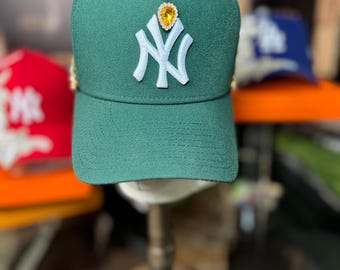 May include: A green baseball cap with a white "NY" logo on the front. The cap is adorned with gold-colored studs around the crown and a large yellow gem at the center. Other caps are visible in the background.