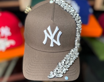 May include: A brown baseball cap with a white "NY" logo on the front. The cap is embellished with a sparkling crystal design along the side. Other caps in red, green, and blue are visible in the background.