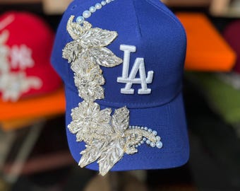 May include: Royal blue baseball cap with the letters "LA" in white. The cap is embellished with silver floral designs and pearl accents. A white button is on top of the cap.