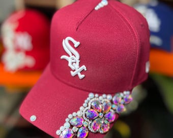 May include: A burgundy baseball cap with the Chicago White Sox logo in white. The brim is adorned with iridescent rhinestones and pearls. Other baseball caps are visible in the background.