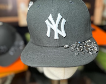 May include: A dark gray baseball cap with the white "NY" logo on the front. The brim is adorned with clear, sparkling rhinestones. The cap is displayed on a mannequin head.