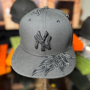 Puede incluir: Gorra de béisbol gris con el logo de los Yankees de Nueva York en pedrería negra. La gorra tiene adornos negros en la visera y la corona. La gorra se exhibe en un soporte de madera.