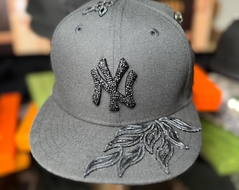 May include: Gray baseball cap featuring the New York Yankees logo in black rhinestones. The cap has black embellishments on the brim and crown. The cap is displayed on a wooden stand.
