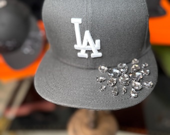May include: A gray baseball cap with a flat brim, featuring the white letters "LA" on the front. The brim is adorned with several clear, sparkling rhinestones. Other black caps are visible in the background.