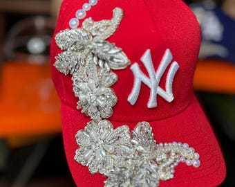 May include: A red baseball cap with a white "NY" logo and elaborate silver and pearl floral embellishments. The cap features a curved brim and a series of small pearls along the side, adding a touch of elegance to the sporty design.