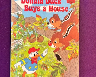 May include: A vintage children's book titled "Donald Duck Buys a House" by Walt Disney Productions. The cover features Donald Duck, Chip and Dale, and colorful illustrations of a house, trees, and foliage. The book has a blue sky background.