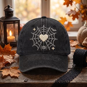 May include: A black baseball cap featuring an embroidered design on the front. The embroidery depicts a grey spider web centered with a cream-colored heart, a small smiling spider, a flying bat, and two four-pointed stars in a light gold color.