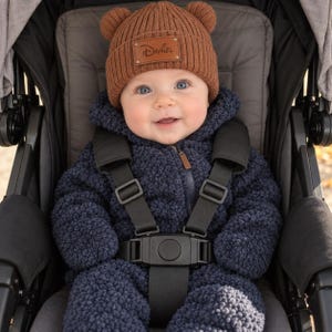 May include: A brown ribbed knit beanie with two small bear ears on top and a rectangular leather patch featuring the name Daniel in a script font, worn by a baby in a navy blue textured fleece hooded jumpsuit with a black five-point harness.
