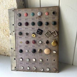 May include: A vintage button display board featuring an assortment of buttons in various colours, shapes, and sizes. The board has a grid layout with numbers and metal edging. Some buttons are round, others square, and some have intricate designs.