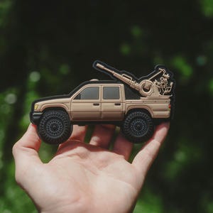 Puede incluir: Parche de camioneta beige con un arma montada en la plataforma. El parche tiene ruedas negras y un borde negro. El fondo es un verde borroso.