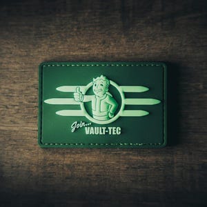 May include: Green rectangular patch featuring the Vault-Tec logo, a smiling cartoon figure giving a thumbs-up. The text "Join... VAULT-TEC" is displayed in white. The patch has a stitched border and is set against a wooden background.