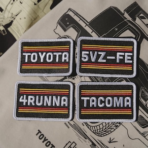 May include: Four rectangular embroidered patches with black backgrounds and white borders. Each patch features text in white, with red and yellow horizontal stripes. The text includes "TOYOTA", "5VZ-FE", "4RUNNA", and "TACOMA".