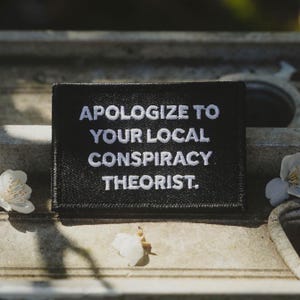 May include: A white ceramic mug with a black handle and the text "But First, Coffee" in a bold, black font. The mug is set against a blurred background, suggesting a cozy setting.