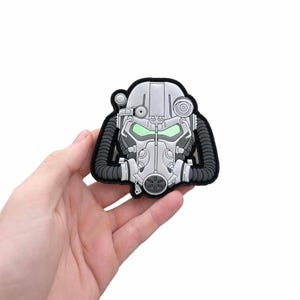 May include: A grey and black patch depicting a detailed illustration of a futuristic helmet with glowing green eye lenses. The patch has a black border and is held in a hand against a white background. The helmet design includes mechanical details.