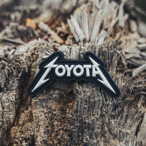 May include: A black and white patch with the word "TOYOTA" in a stylized font. The patch has a black background with white lightning bolt-like accents on each side. The letters are white. The patch is on a rough, wooden surface.