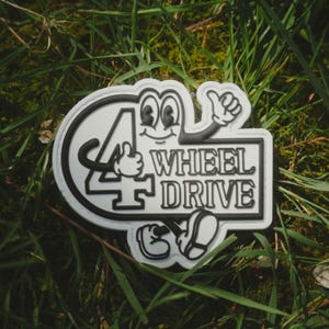 May include: A white and black 4 Wheel Drive badge featuring a cartoon character with a thumbs up gesture. The badge has a black border and is set on a bed of green grass.