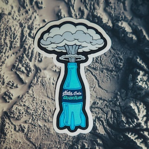 May include: A sticker featuring a blue bottle with the text "Nuka Cola Quantum" and a mushroom cloud design. The sticker has a white border and is set against a textured, gray background.